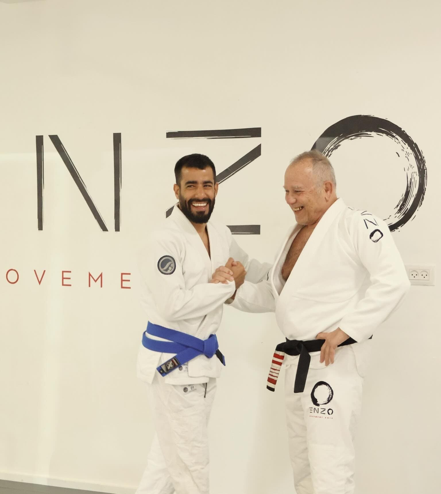 שני מתאמני ג׳יו ג׳יטסו בחליפות לבנות לוחצים ידיים וצוחקים מול קיר הלוגו של אנזו